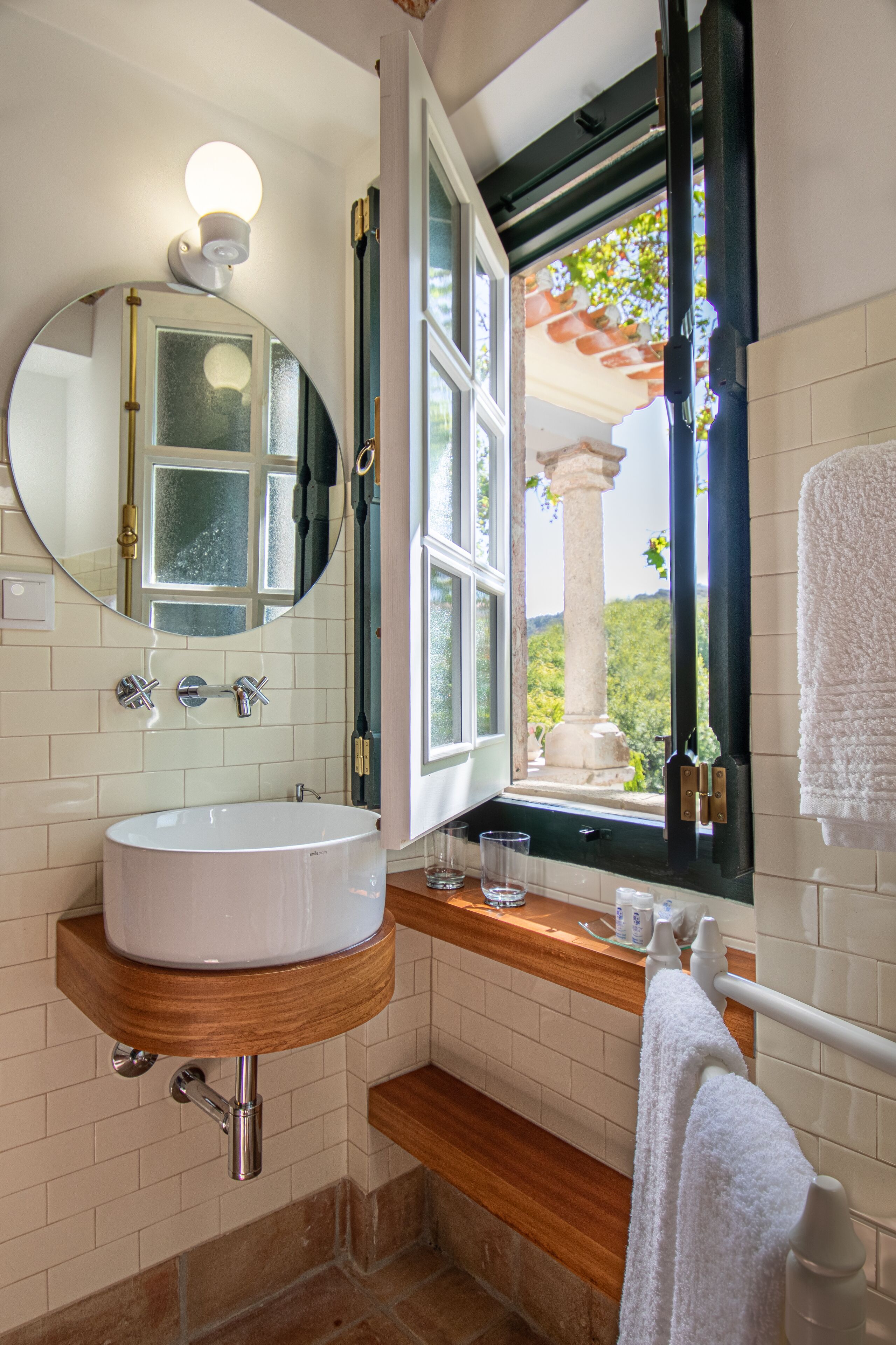 A bathroom with a white sink, a mirror, and a window with an open view of greenery.