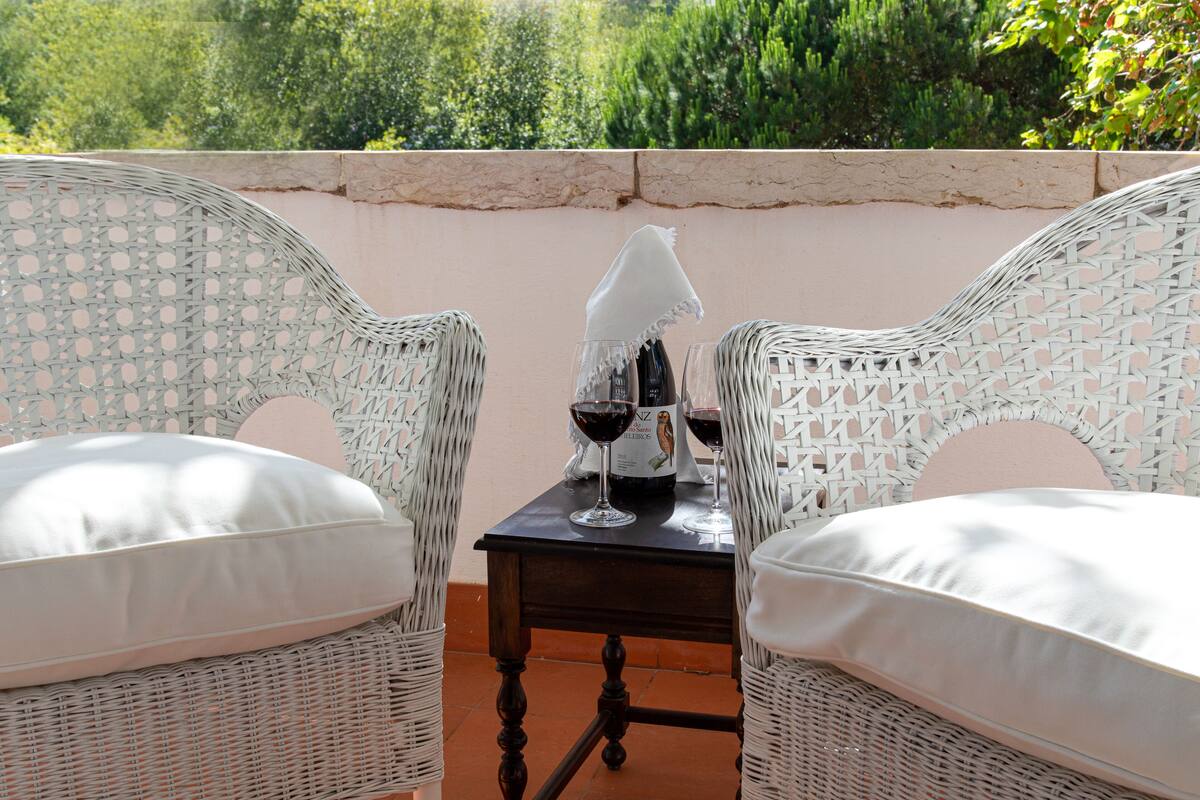 Uma mesa de vime com duas cadeiras, cada uma com uma almofada branca, e uma garrafa de vinho e um copo sobre a mesa.