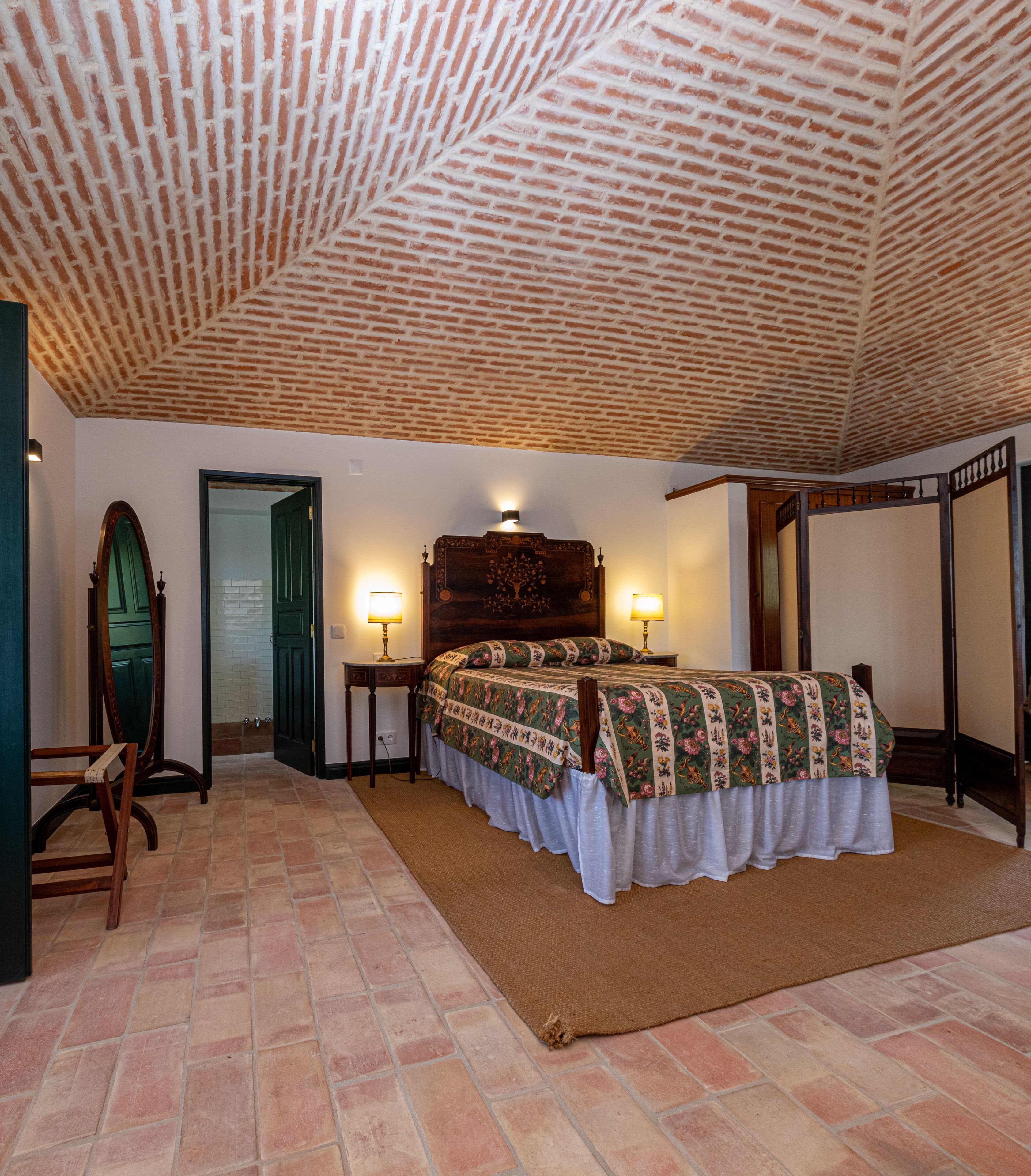 Un dormitorio con techo de ladrillo, una cama con ropa de cama floral, dos lámparas de noche, un cabecero de madera y un espejo grande.
