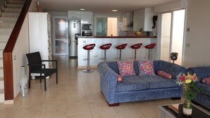 A modern living room with a blue sofa, a black chair, and a coffee table with a vase. A kitchen is visible in the background with a bar stool area. - Nice new Duplex with big terraces, whith very nice seaviews. For your relax (Porís de Abona)