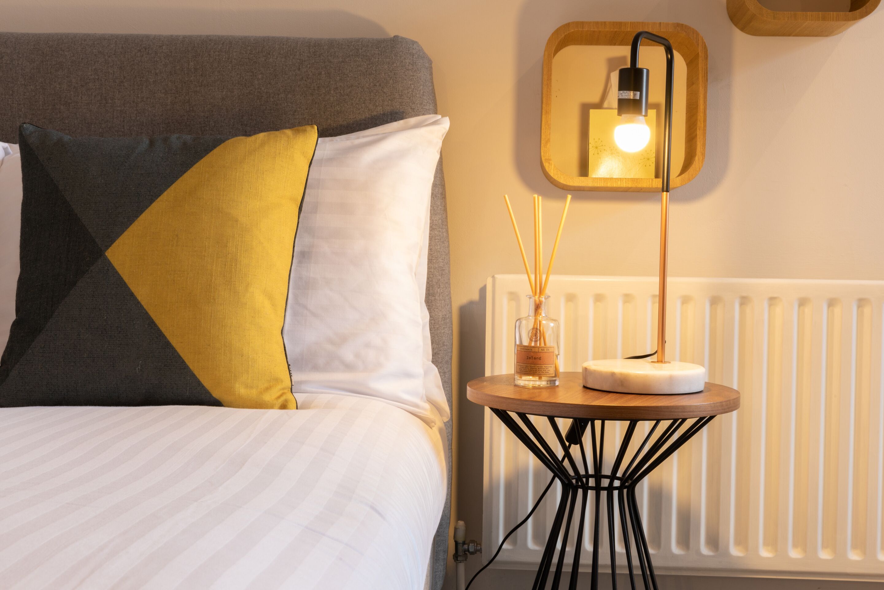 A neatly made bed with a striped white duvet, a grey headboard, and a yellow pillow. A small round table with a white candle and reed diffuser is placed beside the bed.