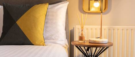 A neatly made bed with a striped white duvet, a grey headboard, and a yellow pillow. A small round table with a white candle and reed diffuser is placed beside the bed.