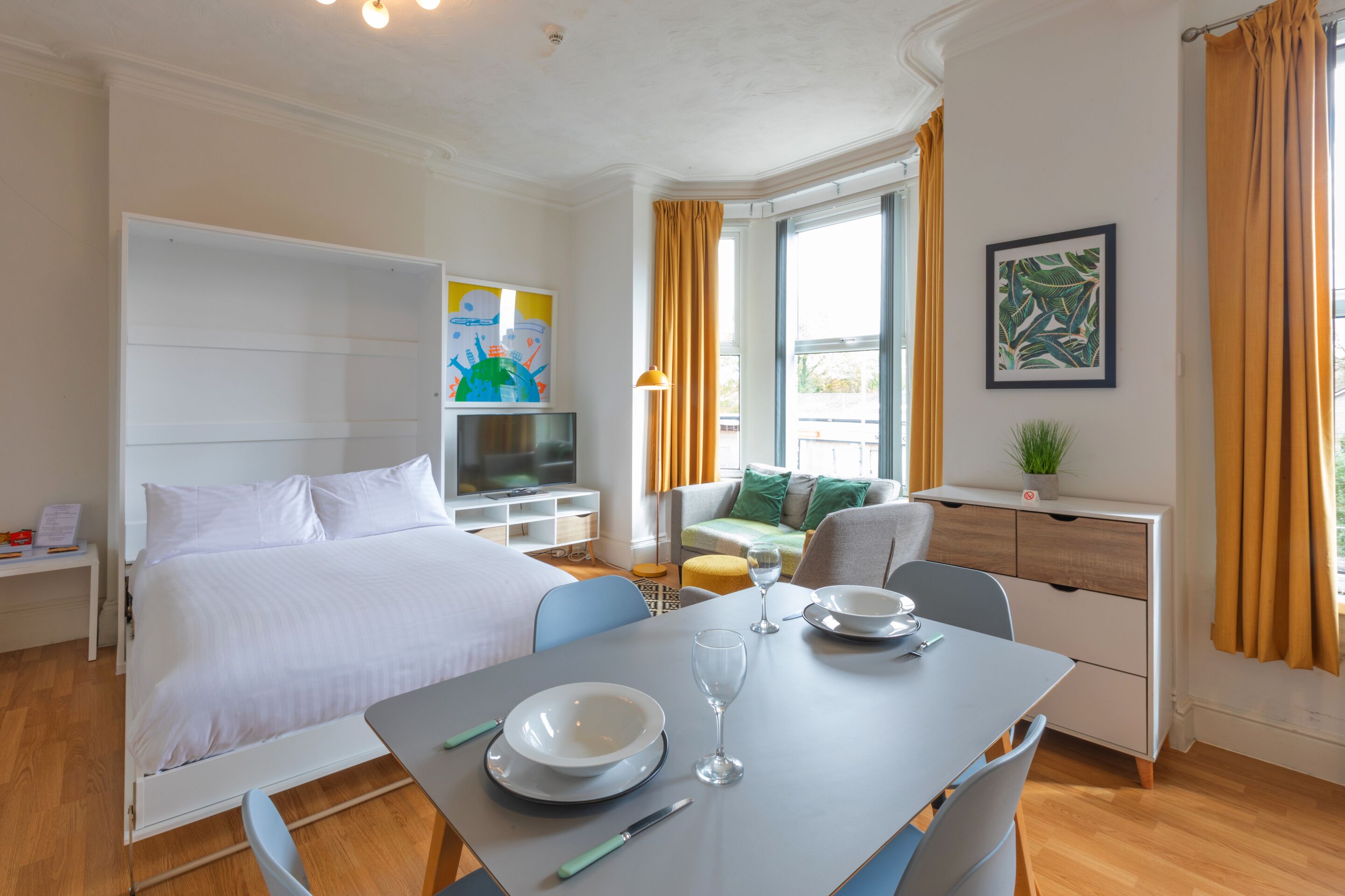 A hotel room with a bed, a TV, a dining table with a glass, a bowl, and a knife.