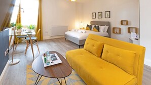 A modern hotel room with a bed, a yellow sofa, a dining table with chairs, and a window with curtains. - Fulwood Heights (Preston)