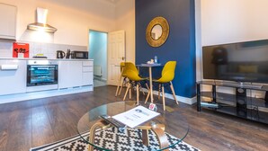 A modern living room with a glass coffee table, a flat-screen TV, and a dining area with yellow chairs. - Fulwood Heights (Preston)