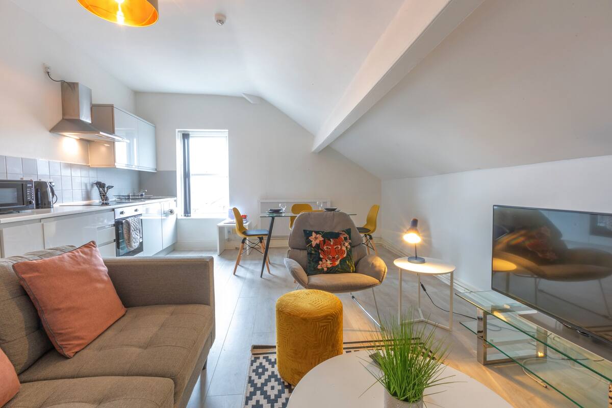 A modern apartment with a kitchenette, a living area with a sofa and ottoman, a dining table with chairs, and a TV.