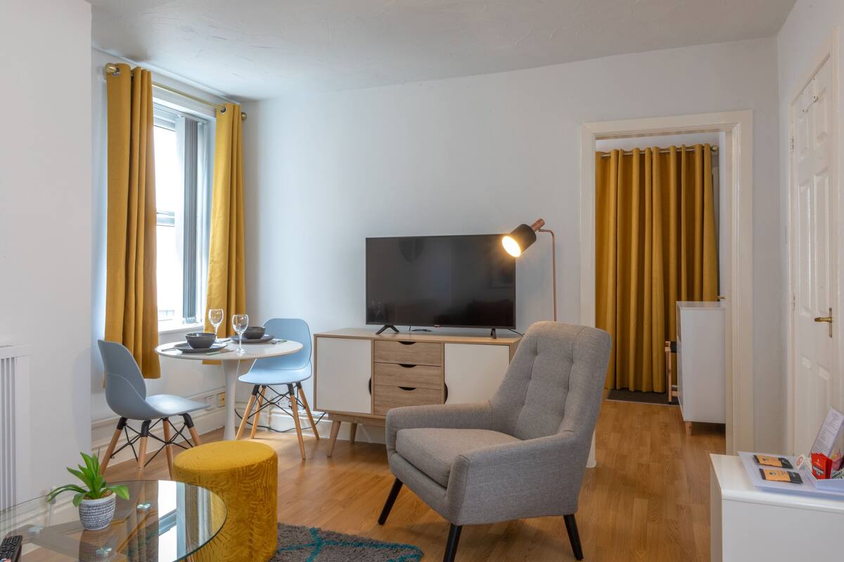 A modern living room with a flat-screen TV, a grey armchair, a small round table, and a glass coffee table.