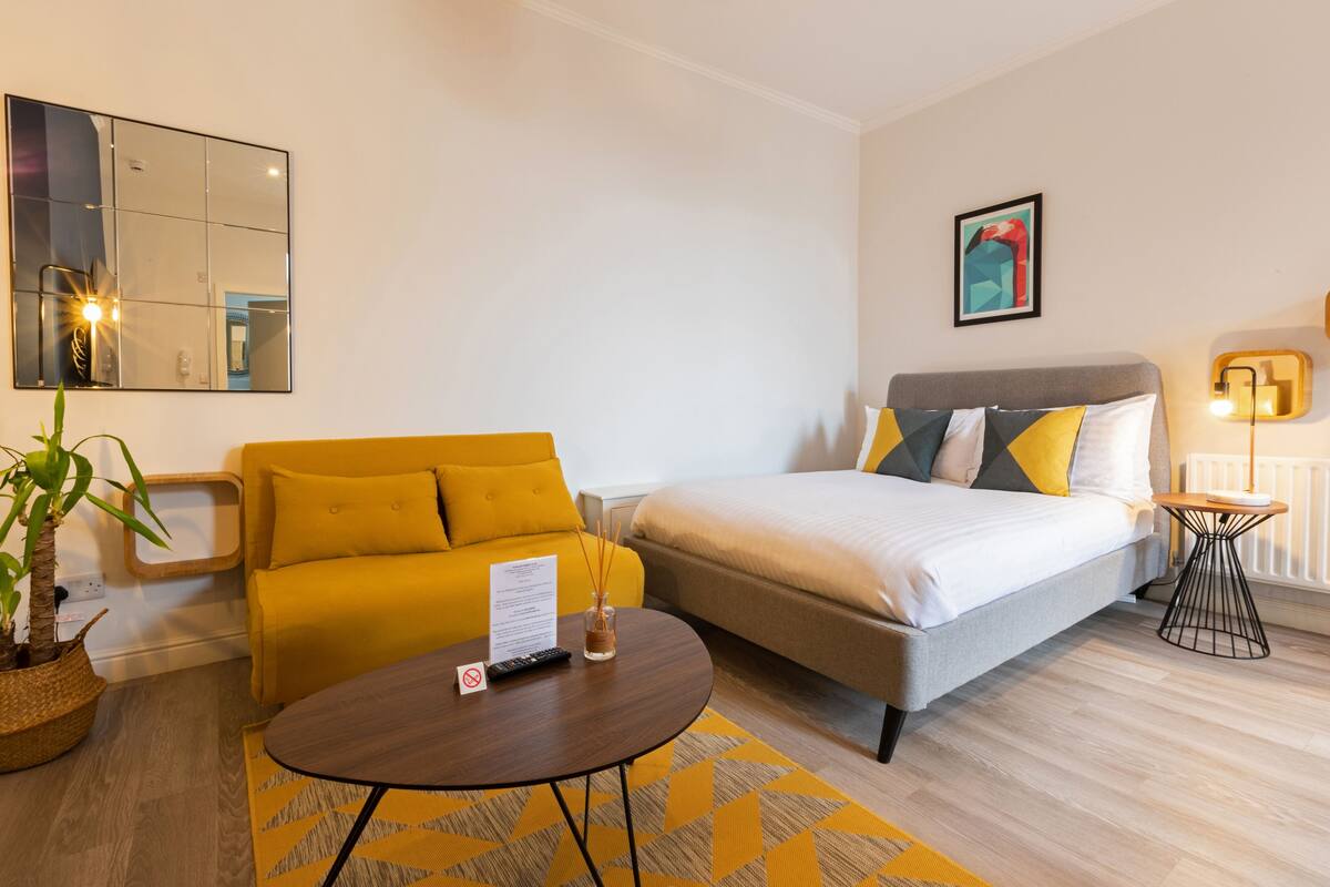 A modern hotel room with a bed, a sofa, a round table, a mirror, and a small table with a lamp.
