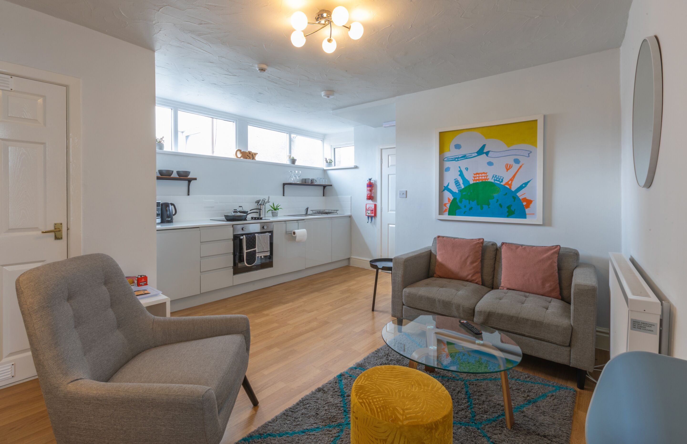 Ein modernes Wohnzimmer mit einem grauen Sessel, einer Couch mit Kissen, einem Glastisch und einem farbenfrohen Teppich. Es gibt eine Küchenzeile mit weißen Schränken, ein Fenster und ein Bild an der Wand.