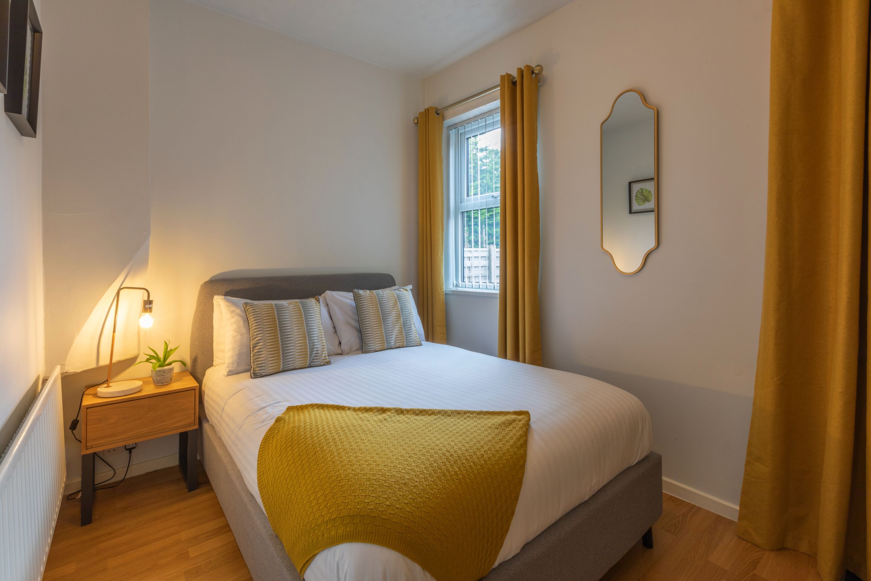 A neatly made bed with a white comforter and a mustard-colored throw blanket, a bedside lamp, and a small potted plant on a wooden nightstand.