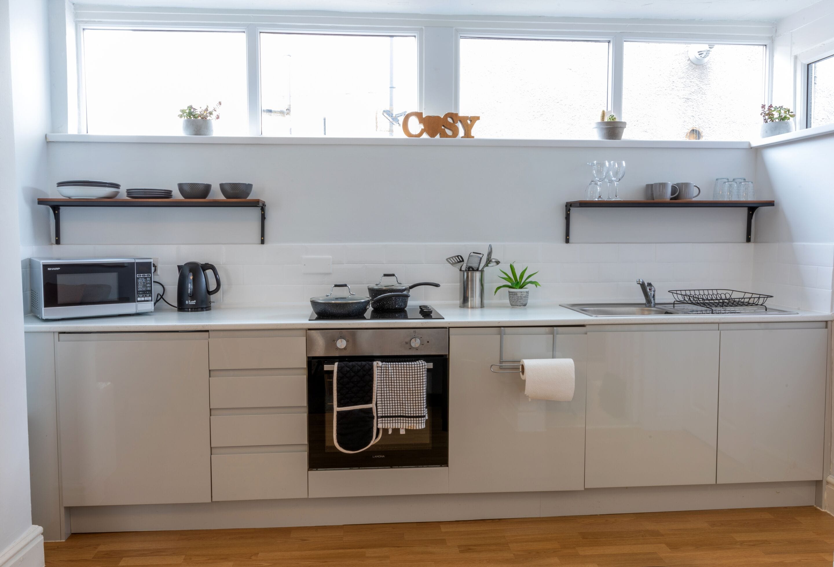 Una cucina moderna con mobili bianchi, forno integrato, microonde e macchina del caffè. Ci sono due finestre sopra il lavello e uno scaffale con ciotole e bicchieri.