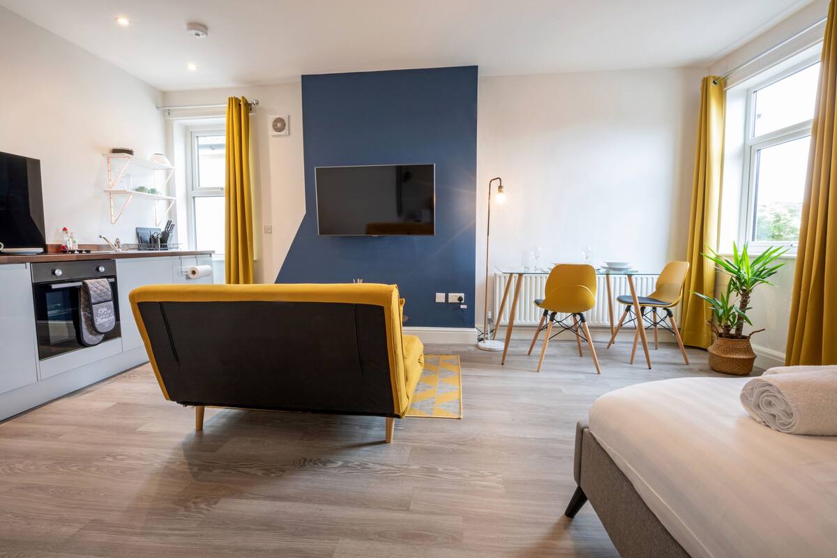 A modern hotel room with a bed, a sofa, a TV mounted on the wall, a dining area with chairs, and a kitchen area.