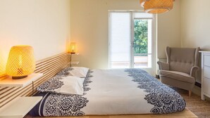 Ein Schlafzimmer mit Bett, Nachttischlampen, einem Sessel und einem Fenster mit Blick auf Grünflächen.