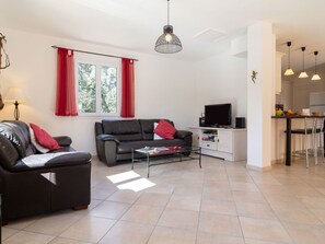 Een moderne woonkamer met een bank, een salontafel en een televisie. Er is een keukenhoek met een koelkast en een eettafel.