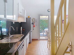 Una cocina moderna con un lavamanos, armarios y un refrigerador. Se ve un comedor con sillas y una mesa a través de una puerta abierta.