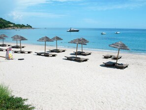 A beach with sun loungers and thatched umbrellas, a calm sea, and boats in the distance.