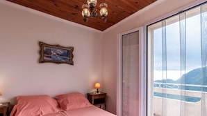 A bedroom with a bed, a nightstand, a lamp, a framed picture, and a sliding glass door leading to a balcony with a view of mountains.