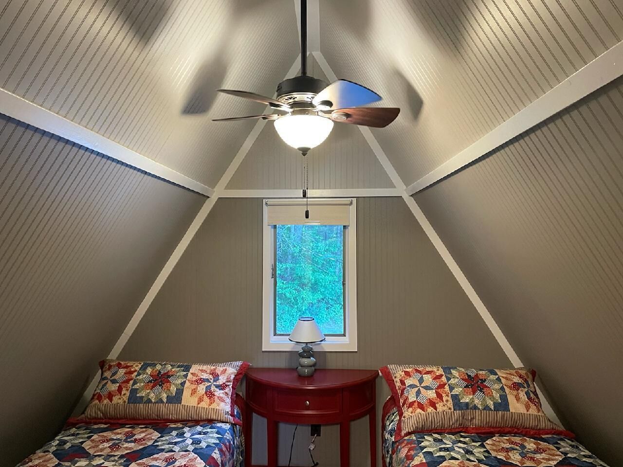 Una camera da letto con due letti, una ventola a soffitto, una finestra vista alberi e un piccolo tavolo con una lampada.