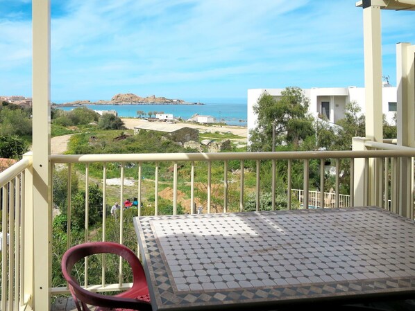 A balcony with a tiled floor and a red chair, overlooking a coastal area with buildings and a clear sky.