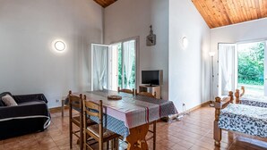 A spacious room with a wooden ceiling, a dining table with chairs, a sofa, a TV, and large windows offering a view of greenery. - Cosy apartment in villa for 6 guests with TV, terrace and pets allowed (Coggia)