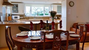 Un coin repas avec une table en bois dressée pour quatre personnes, une cuisine en arrière-plan et une horloge au mur.