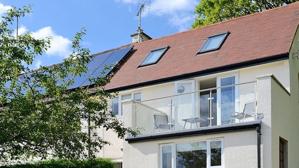 Une maison à deux étages dotée d’un balcon, de panneaux solaires et d’un jardin.