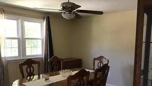 Comedor con una mesa redonda de madera, seis sillas, un ventilador de techo y una ventana con cortinas.