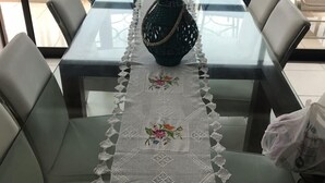 A dining table with a glass top, surrounded by cushioned chairs, a decorative woven basket, and a lace table runner with floral embroidery.