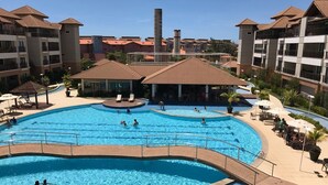 A large outdoor swimming pool with multiple sections, surrounded by buildings with balconies and a clear sky.
