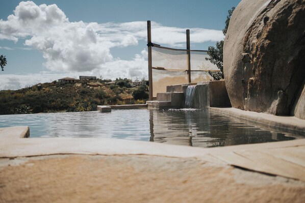 Una piscina con una cascada, escalones de piedra y una gran roca.