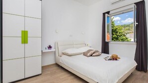 A bedroom with a bed, a wardrobe, a window with curtains, and a view of greenery outside. - Apartments St.Thomas - Deluxe Two-Bedroom Apartment (Dubrovnik)