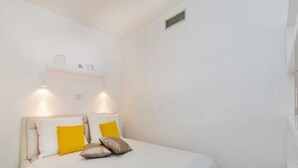 A bedroom with a bed, white walls, a vent, and a shelf with items. - Apartments St.Thomas - Deluxe Studio with Terrace (Dubrovnik)