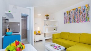 A modern living room with a yellow sofa, a white coffee table, and a fruit bowl. There is a kitchen area with a blue countertop and a white cabinet. A colorful abstract painting is on the wall. - Apartments St.Thomas - Deluxe Studio with Terrace (Dubrovnik)