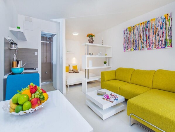 A modern living room with a yellow sofa, a white coffee table, and a fruit bowl. There is a kitchen area with a blue countertop and a white cabinet. A colorful abstract painting is on the wall.