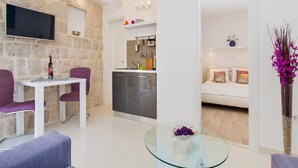 Ein modernes Wohnzimmer mit einer Wand aus Stein, einem Flachbildfernseher, einem Essbereich mit violetten Stühlen, einer Küche mit Einbauofen und einer gemütlichen Sitzecke mit weißem Sofa.