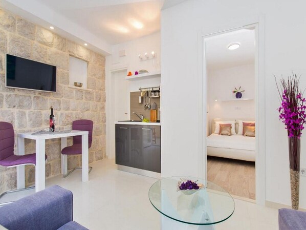 Uma sala de estar moderna com parede de destaque em pedra, uma TV de tela plana, uma área de jantar com cadeiras roxas, uma cozinha com forno embutido e um espaço de estar aconchegante com um sofá branco.