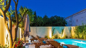 Un espace aménagé au bord de la piscine, avec des chaises longues en bois et une petite table, entouré de verdure et avec un bâtiment en arrière-plan.
