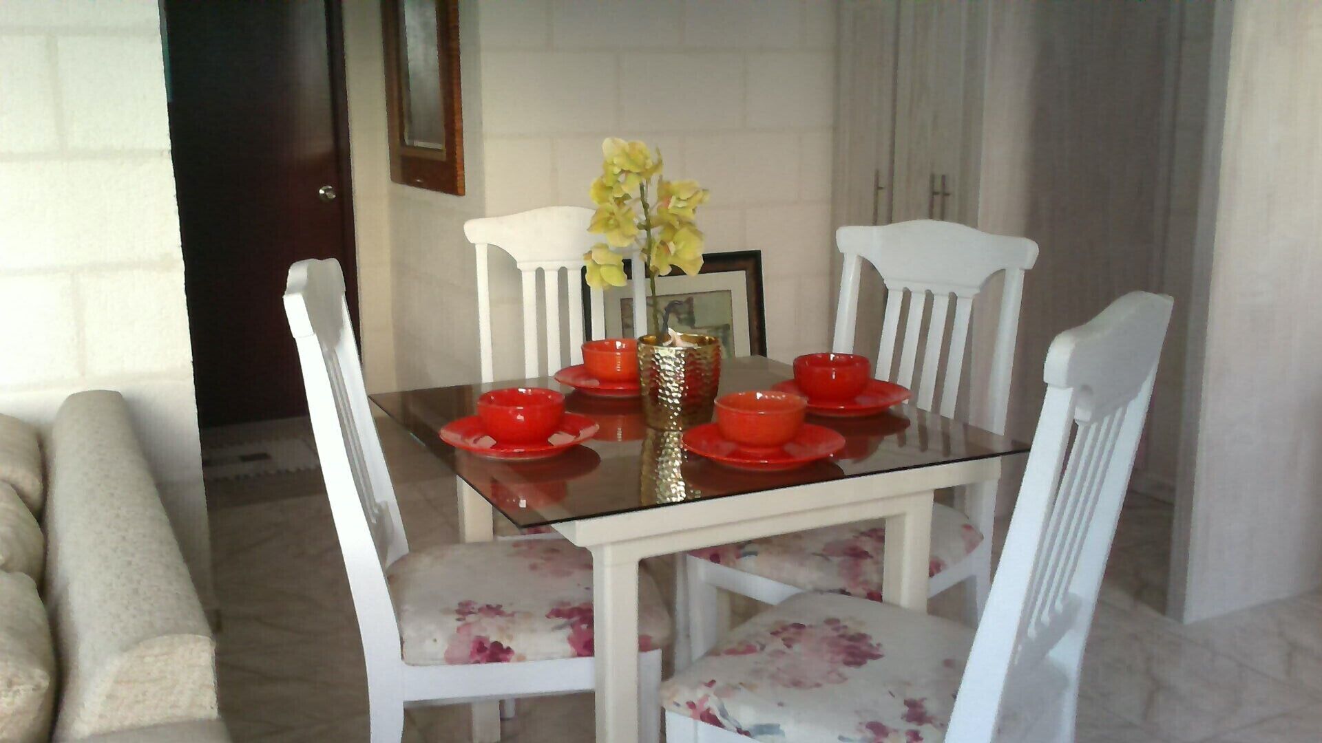 Een eethoek met een glazen tafel, witte stoelen met bloemrijk beklede kussens, en rode kommen en een vaas met gele bloemen.