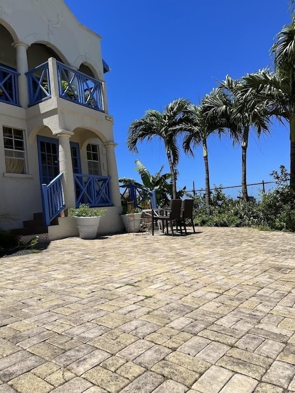 A building with a white exterior and blue accents, a paved patio with a table and chairs, and palm trees in the background.