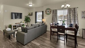 Un salon moderne avec un canapé gris, une table à manger avec des chaises, un téléviseur fixé au mur et une horloge.