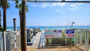 Acceso a la playa con palmeras, una pasarela de madera y varios carteles de advertencia.