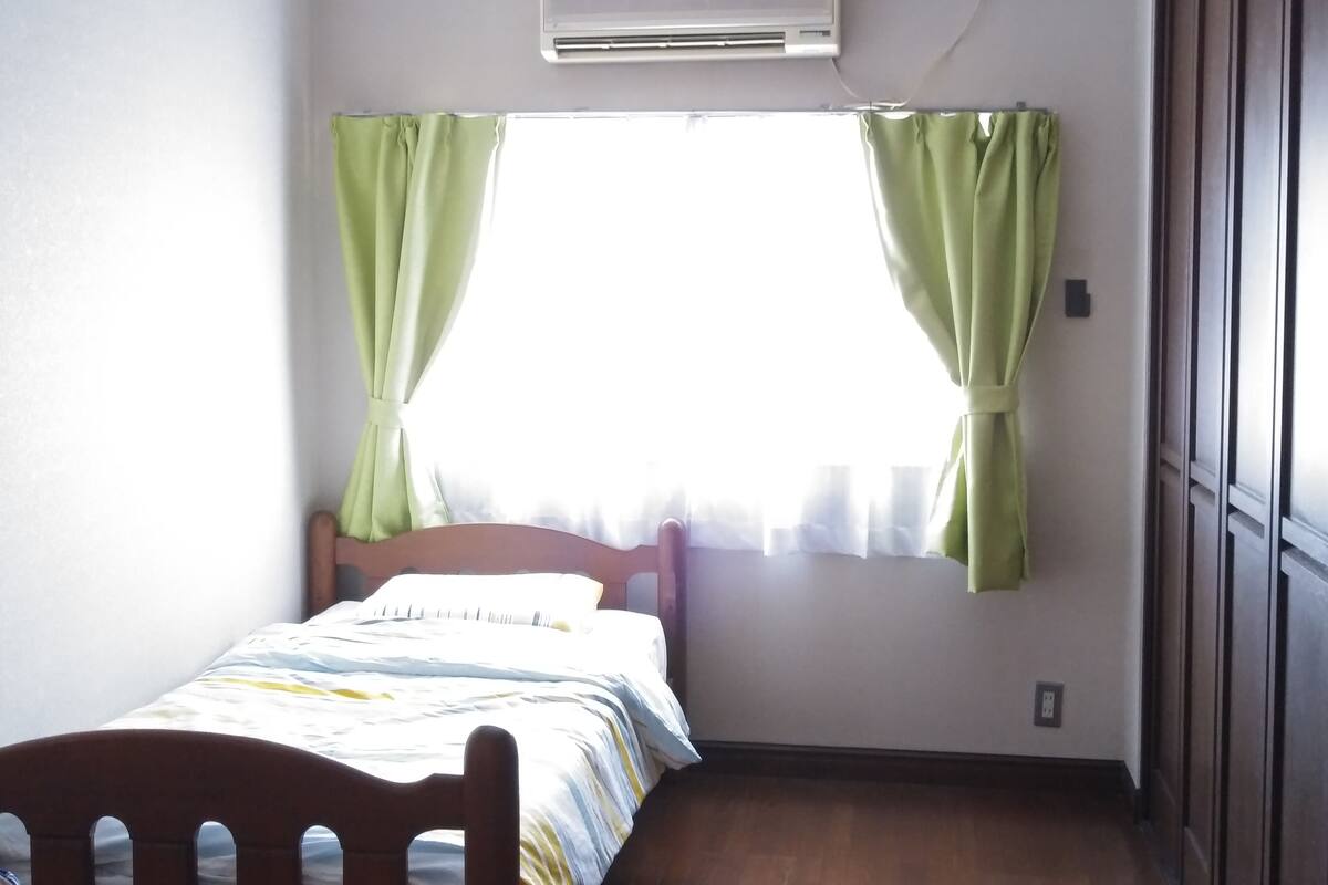 A small, well-lit room with a bed, a window with green curtains, and an air conditioning unit on the wall.