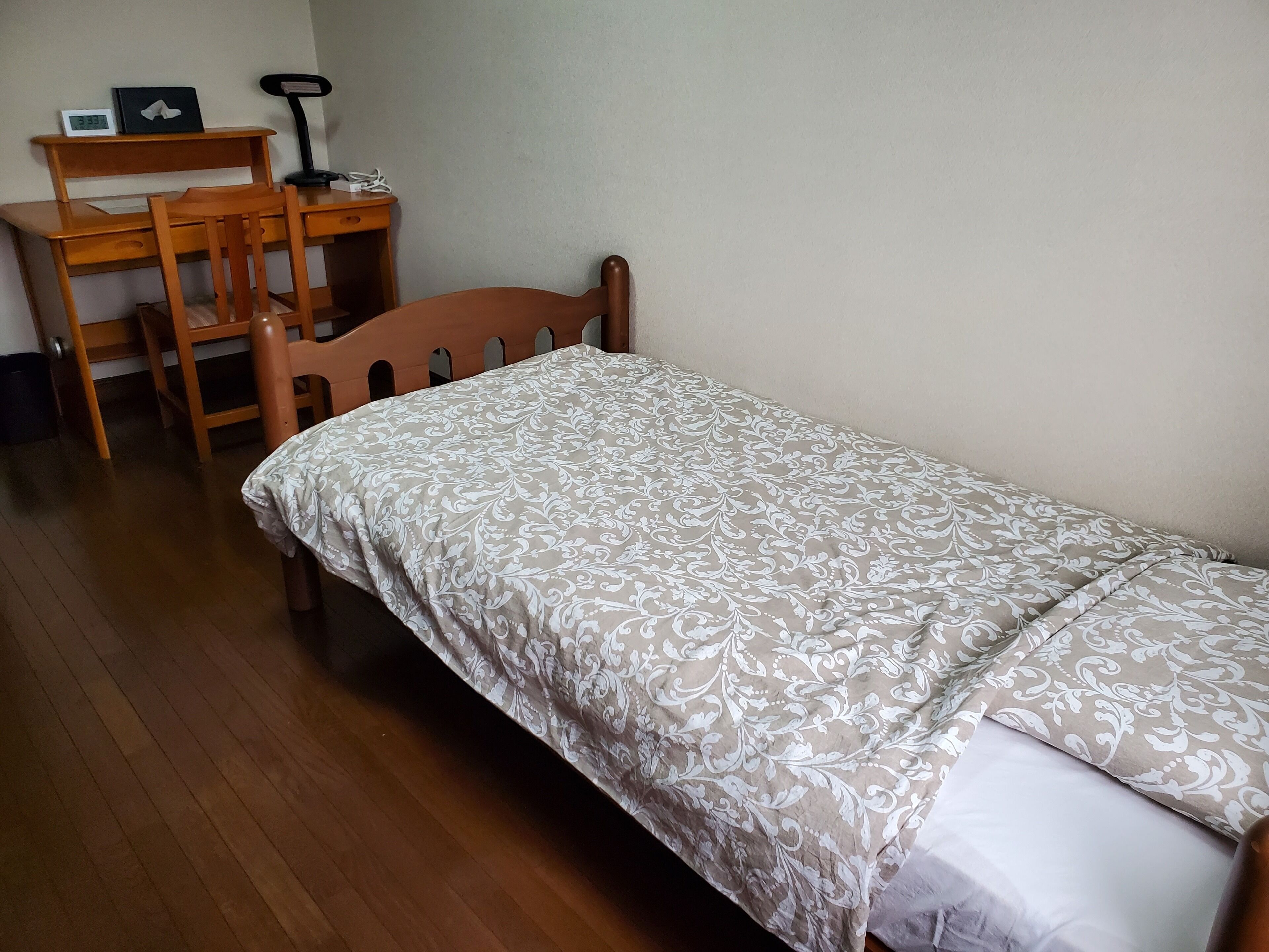 A small, single-bed room with a wooden bed frame and a patterned bedspread. There is a wooden desk with a chair and a clock on it.