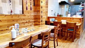 Un interior de madera con una mesa larga y sillas, un mostrador con una botella blanca, y una pizarra con el menú.