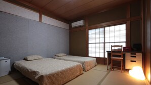 Una habitación japonesa tradicional con suelo de tatami, dos camas con ropa de cama a juego, un escritorio con silla y una ventana con persianas.