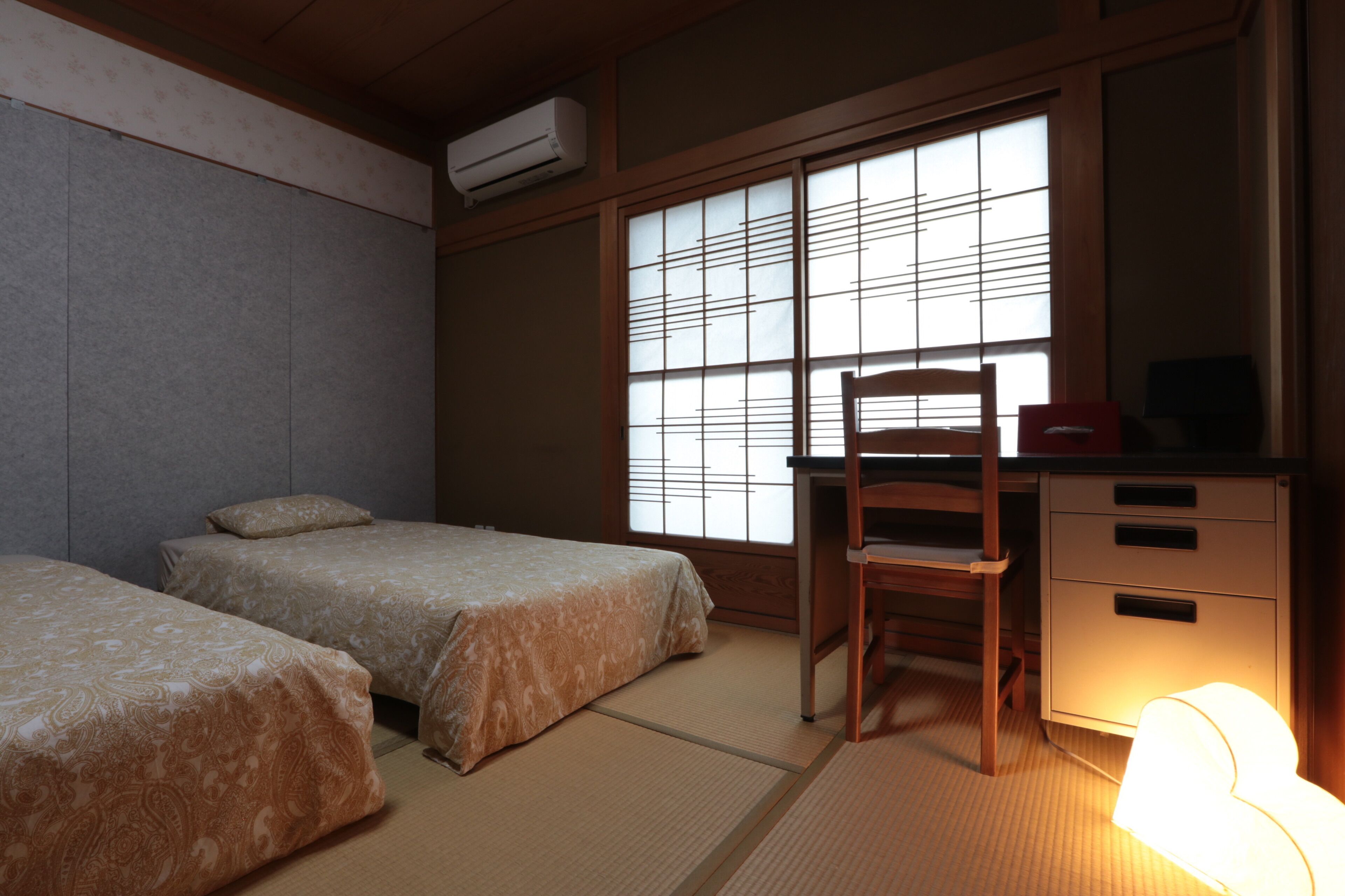A traditional Japanese room with tatami flooring, two beds, a desk, and a chair.