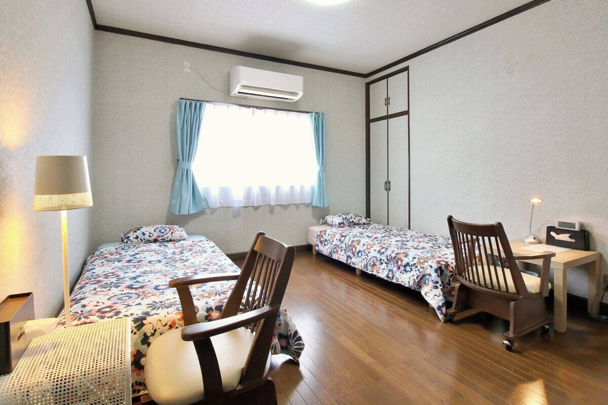 A room with two beds, a desk, a chair, an air conditioner, and a window with curtains.