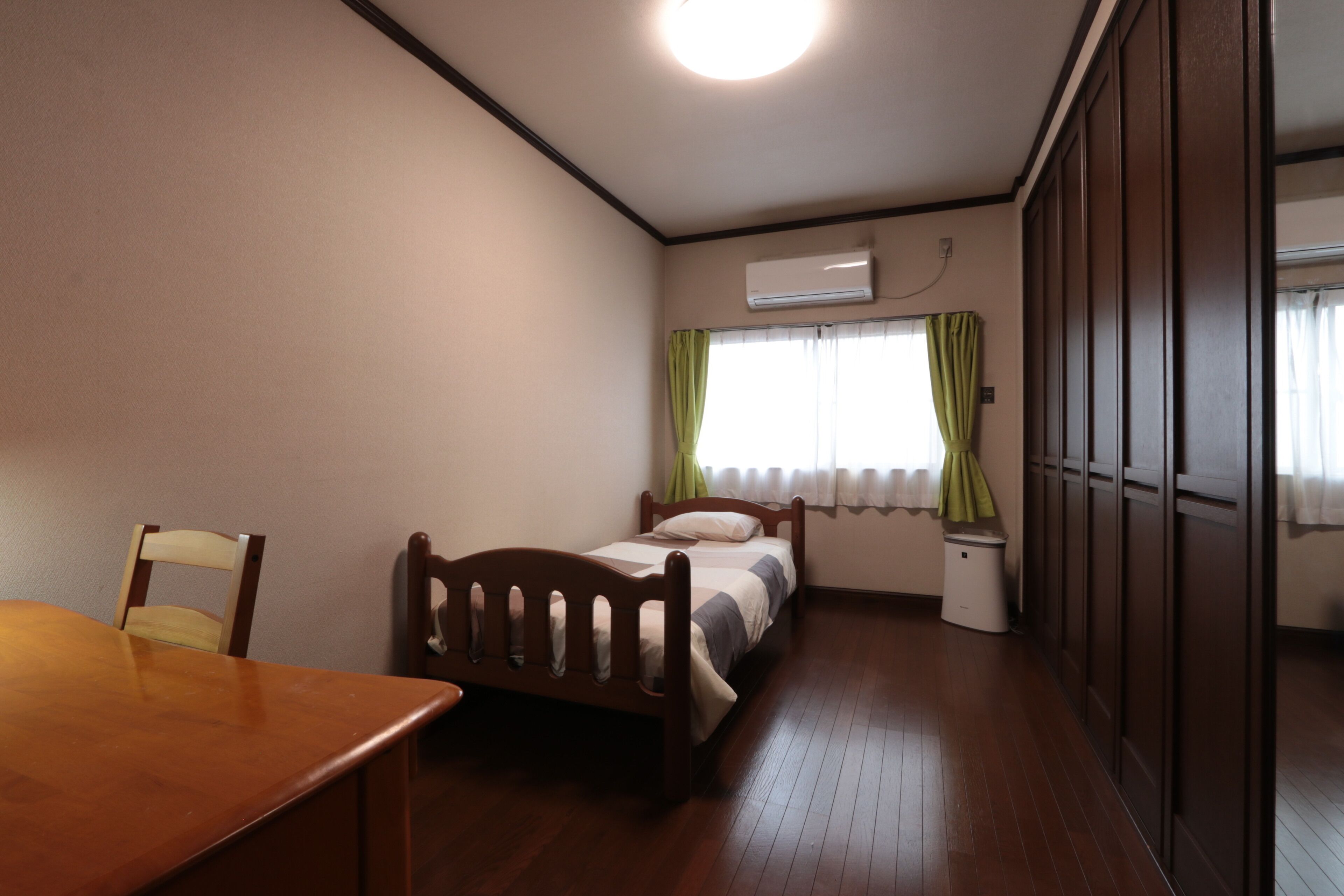 Un dormitorio con cama de madera, una mesita de noche pequeña, una silla, una ventana con cortinas y un equipo de aire acondicionado.