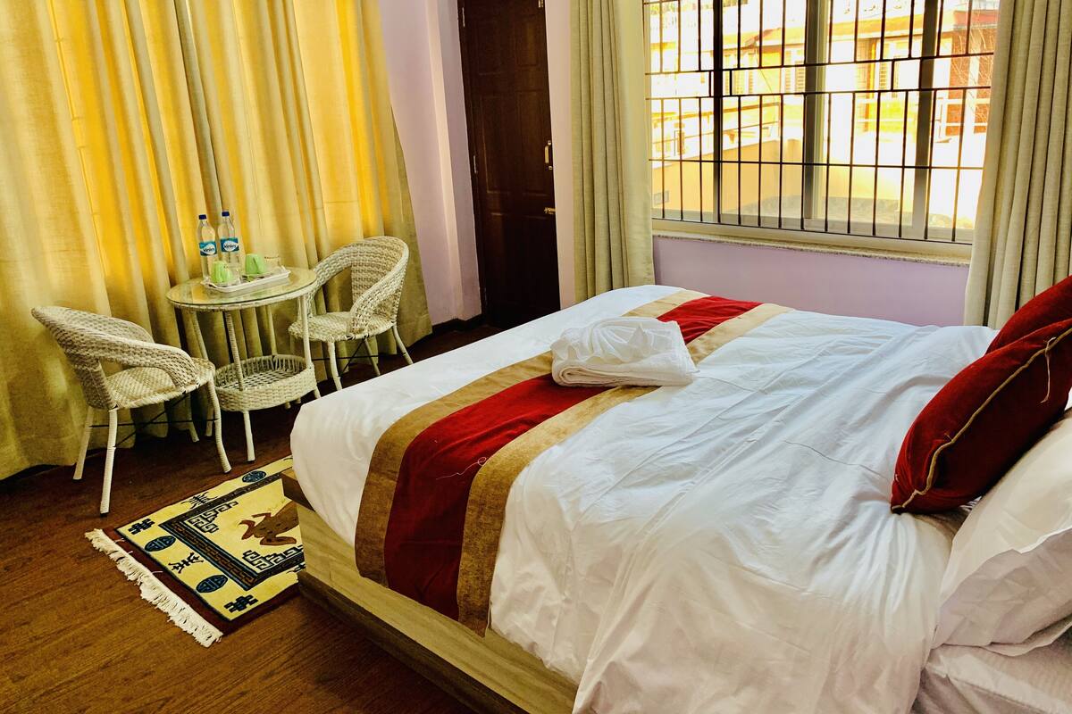 Una habitación de hotel con una cama, una mesita con sillas, una ventana con rejas y cortinas.
