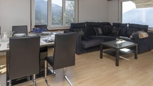A modern living room with a dark sofa, a coffee table, and a dining area with chairs and a table set for two. Large windows provide a view of the outdoors. - Maison Le Fontany by Interhome (Crans-Montana)